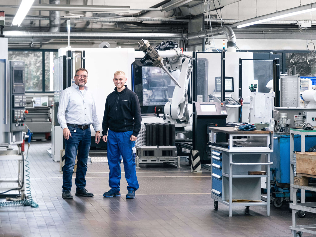 Zwei Personen stehen in einer Produktionshalle. Links ein Mann in einem weißen Hemd, rechts ein Mann in blauer Arbeitskleidung. Im Hintergrund sind Maschinen und ein Roboter sichtbar.