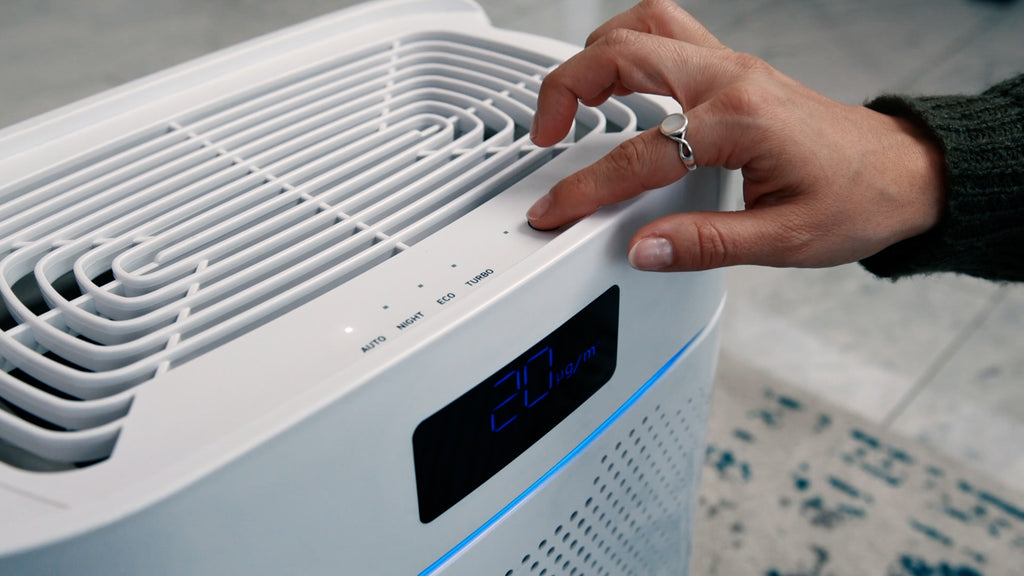 Vorschaubild zum Video über den Luftreiniger IDEAL AP25 – der Zoom liegt ausschnitthaft auf dem Geräteoberteil. Zu sehen sind die oberen Luftauslässe, die Touch-Bedienelemente sowie seitlich vorne ein Display mit blauer PM 2.5-Anzeige. Eine Hand im Bild betätigt die Ventilator-Taste.