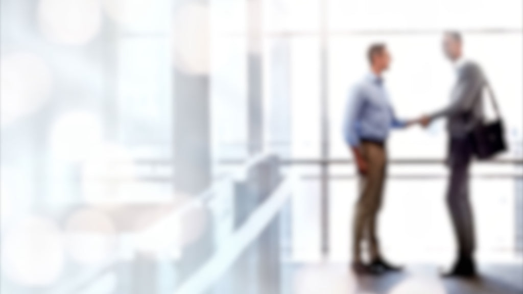 Zwei Männer schütteln sich die Hände in einem modernen Gebäude mit großen Fenstern. Einer trägt ein Hemd und eine Hose, der andere ein Hemd und eine Tasche.