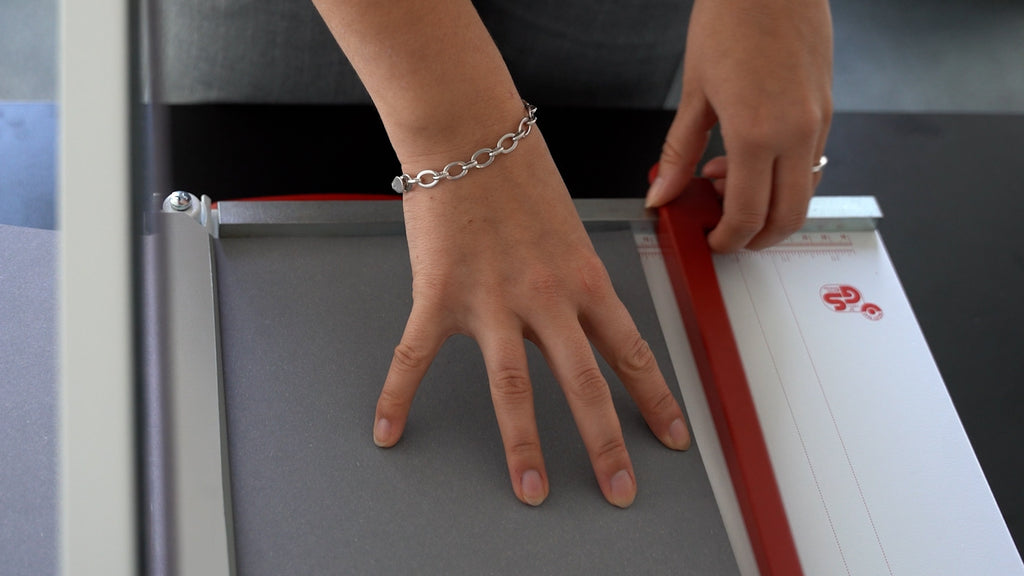 Szenenbild des Videos zur Einstellung des Rückanschlags des IDEAL Hebelschneiders 1046 auf das gewünschte Maß: Der Zoom liegt auf dem Schneidetisch mit mm-/Zoll-Maßskala. Eine Hand justiert den Rückanschlag, während die andere auf einen grauen Papierbogen drückt.