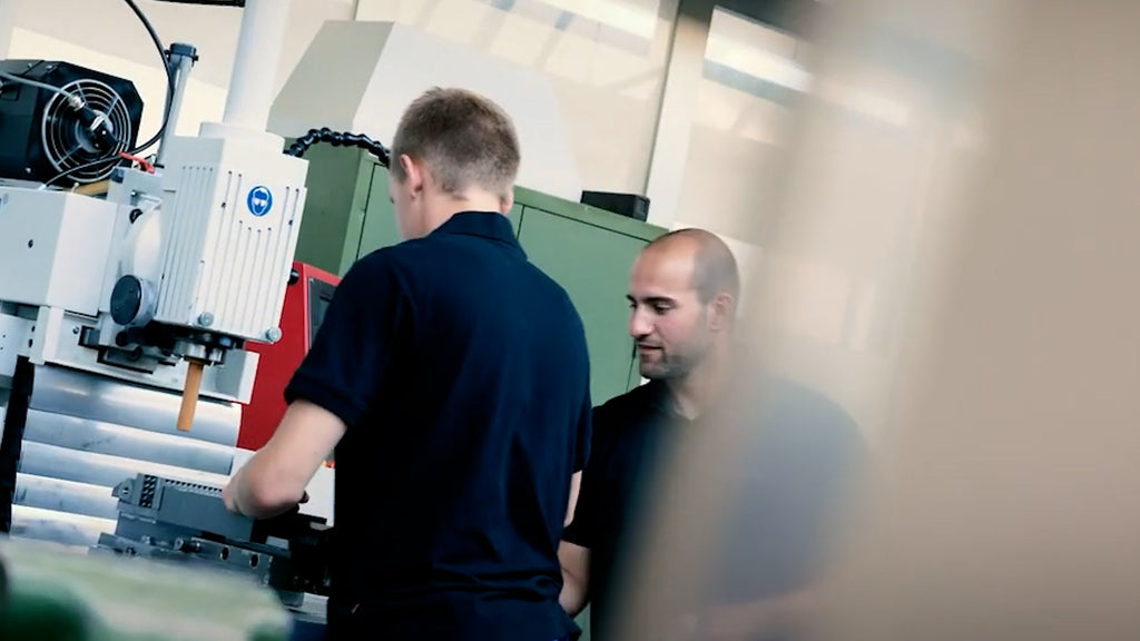 Zwei Personen in Arbeitskleidung  befinden sich in einer Maschinenwerkstatt. Eine bedient eine Maschine, während die andere daneben steht. 