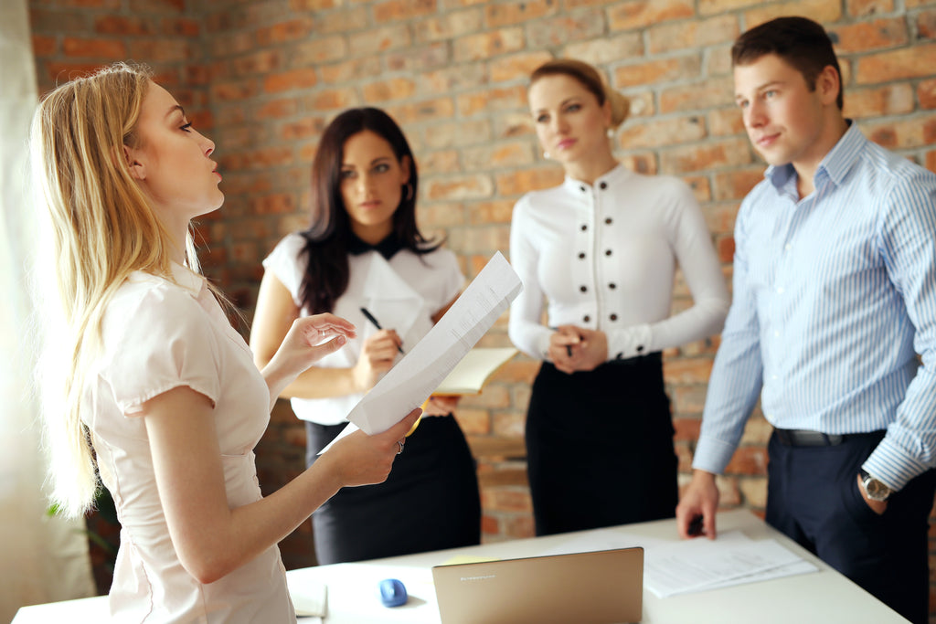 Eine Gruppe von 4 jungen Leuten in eleganter Office-Kleidung steht um einen Schreibtisch in einem Büro mit rohen Ziegelwänden und diskutiert. Eine Person spricht gerade, die anderen hören aufmerksam und etwas skeptisch zu.